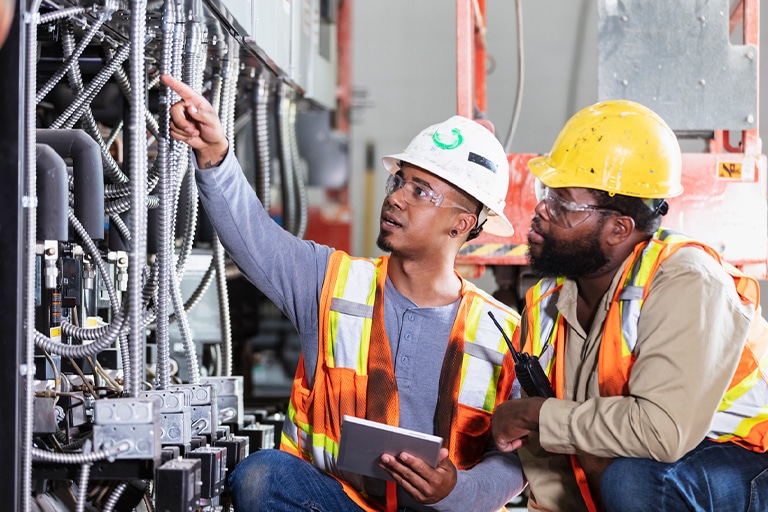 Two technicians examining electrical panels, wearing safety helmets and vests in an industrial setting.